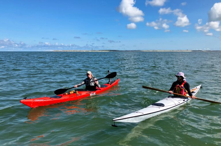 Twee kajaks op de zee tussen Ameland en Terschelling met het Amelander stand, de duinen en de vuurtoren als een dun streepje op de achtergrond.
