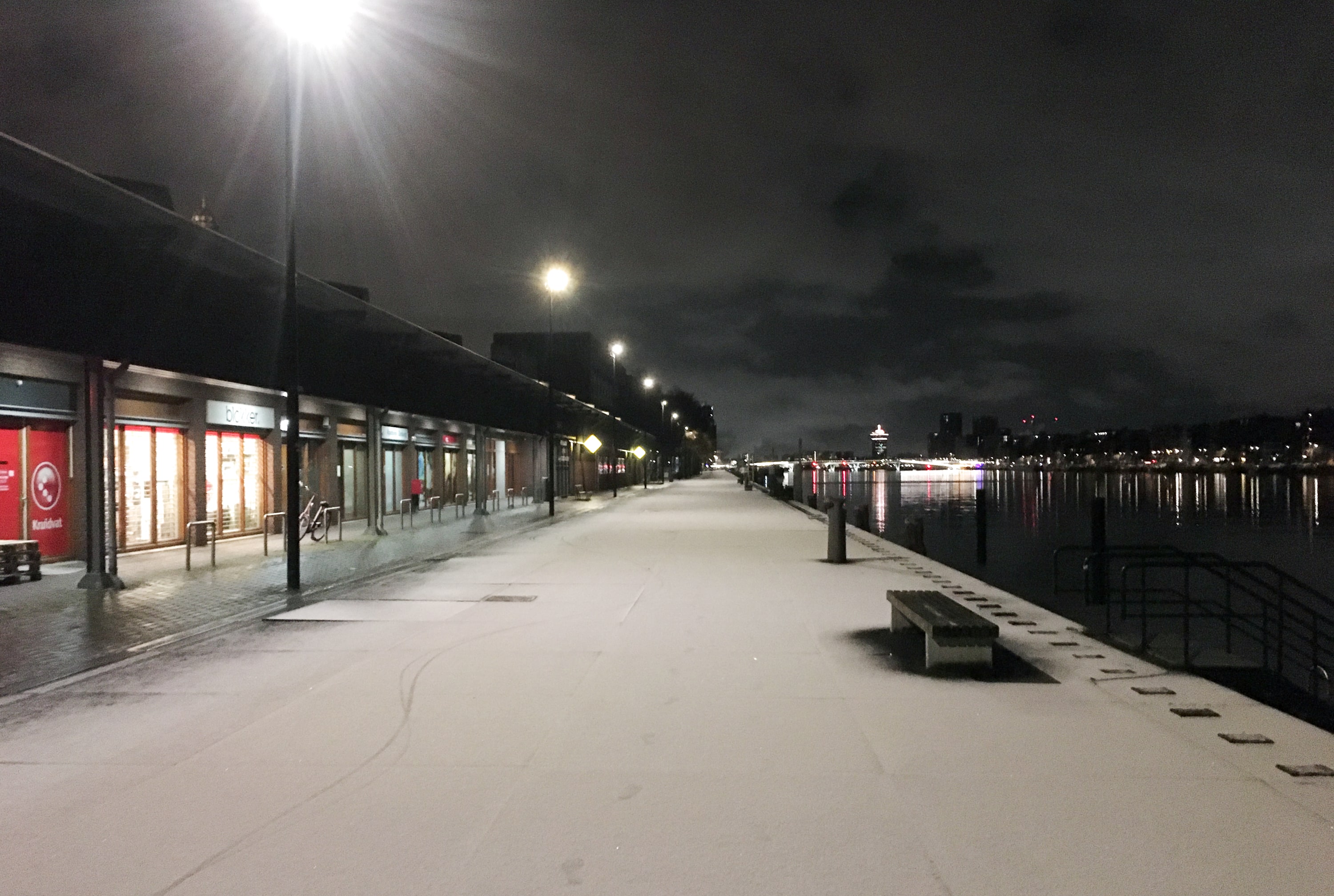 De Veemkade in Amsterdam met een laagje witte sneeuw in een verder donkere wereld. Met links lantarenpalen die als zonnetjes schijnen en rechts het donkere spiegelende water.