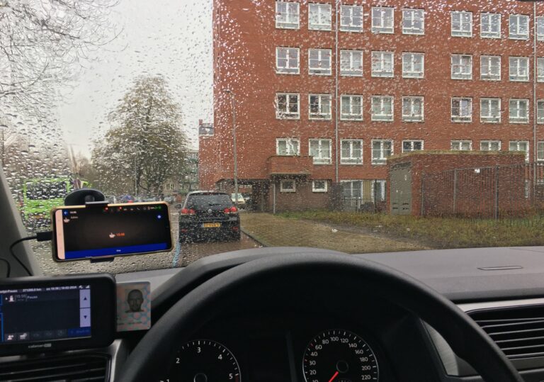Uitzicht vanuit mijn voorruit van een wat saai flatgebouw van 5 verdiepingen en wat geparkeerde auto's. De regendruppels op het voorruit maken er een beetje mozaïek van.