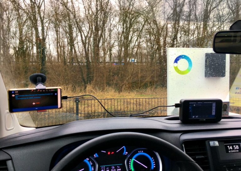 Uitzicht vanuit de voorruit van de auto. Regendruppels op het raam en een kale bomenrij erachter. Daarachter een hint van de snelweg. De auto staat bij een snellaadpunt.