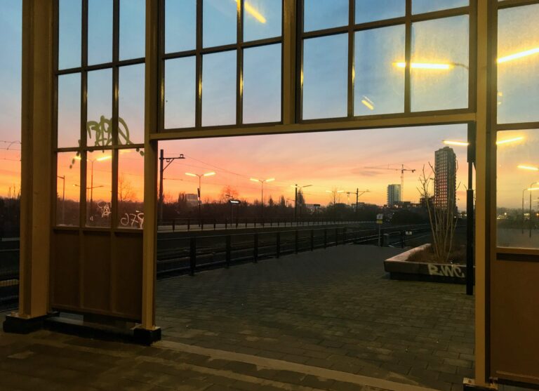 Een foto van de zonsopkomst bij het Amstelstation in Amsterdam. De lucht kleurt geel, oranjerood en bovenin al blauw. En het zonlicht weerkaatst in de ramen van het flatgebouw op de achtergrond.