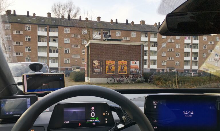 Sta geparkeerd aan de rand van de weg met uitzicht op een elektriciteitsgebouwtje van rode baksteen met daarachter een appartementencomplex van 4 hoog ook opgetrokken uit rode baksteen met witte balkonnetjes. 