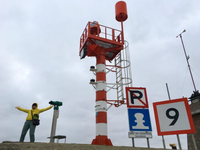 Els op het havenhoofd bij Oudeschild. Lekker uitwaaien, armen wijd. Samen met een verrekijker op een paal, twee aanwijsborden voor varende mensen, een windroos en het havenlicht, een grote rood met witte paal met daarop een plateau met een hek erom.