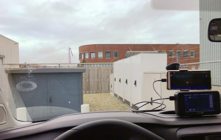 Uitzicht vanuit mijn voorruit van de auto terwijl het aan de snellader staat. Heel on inspirerend. Wat elekctra kasten in grijs en wit, een schutting links en rechts in de hoekjes stukjes van loodsen en achter de schutting een saai plat rode bakstenen kantoorgebouw. En om de dit allemaal nog wat kracht bij te zetten een grijze lucht met wat lichte regenwolken.