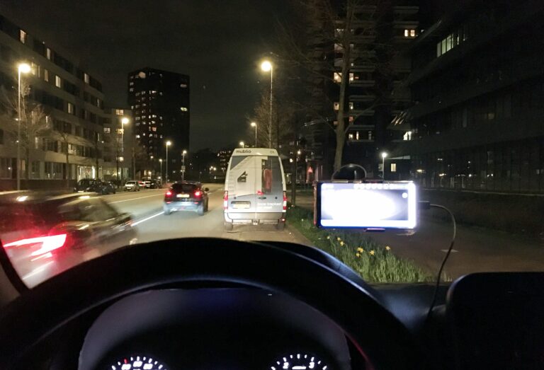 Uitzicht vanuit mijn voorruit van de auto. Het is donker. Twee auto's rijden voorbij en een witte bus met opdruk staat voor mij geparkeerd. Ik kijk uit op twee flatgebouwen