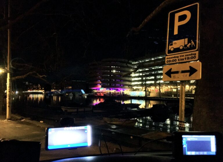 Uitzicht vanuit een autoraam in het donker. Op de voorgrond rechts een blauw bord over laden en lossen en op de achtergrond bootjes de gracht en daarachter de verlichte flat met 8 verdiepingen waar auto's wonen. Met zijn klassieke rondingen waardoor auto's naar de volgende verdiepingen kunnen komen.