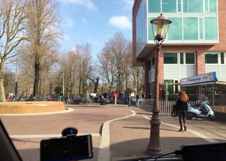 Uitzicht vanuit mijn autoraam met een parkeerplek bij het OLVG in Amsterdam Oost. Heb zie een kleine rotonde en de ingang rechts naar de parkeergarage en een stukje van het ziekenhuisgebouw. Achter de rotonde fietsen en daarachter het Oosterpark. Het is een mooie zonnige dag.