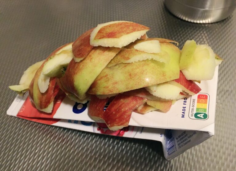 Op een metalen keukenblad ligt een uitgeknepen alpro yoghurt pak (mild & creamy zonder suiker) met daar bovenop de schillen en klokhuizen van twee appels. 