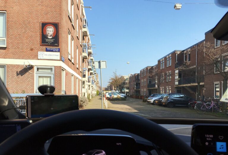 Uitzicht vanuit mijn autoraam op de straat met aan beide kanten appartementen met 3 lagen. Zijn blokkendozen. Zon schijnt op de linkerkant van de straat en de lucht is blauw.
