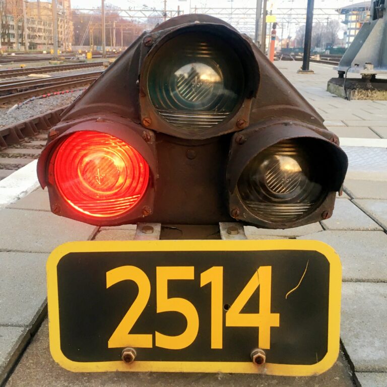 Stoplicht bij een treistation. Bestaat uit 3 lampen in een driehoek. Links is rood en boven is groen. Het geeft de treinmachinist aan wanneer het veilig is te vertrekken. Onderaan een zwart bord met een gele rand met het nummer 2514, ook in geel.