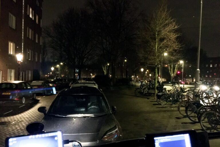 Uitzicht vanuit de voorruit van een autobus. Het is donker. Het enige licht is van de lantaarnpalen. Rechtvoor een aantal geparkeerde auto's. Rechts een tiental geparkeerde fietsen. Daartussen de stoep. Achter wat kale bomen