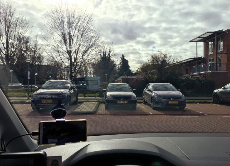 Foto vanuit mijn auto voorruit. Recht naar de zon toe (niet zichtbaar, maar voelbaar). De lucht is vol met watten wolken. Voor me drie geparkeerde auto's met de neuzen naar mij toe. Daarachter wat bomen, struiken en huizen. 