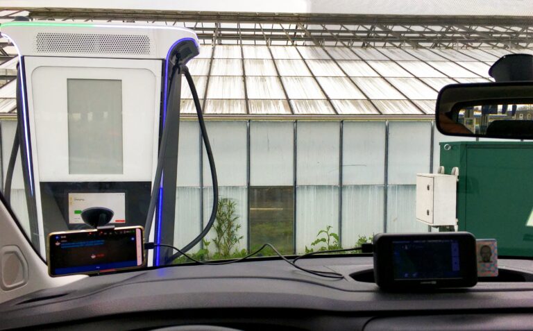 Uitzicht vanuit de voorruit bij een tankstation. Auto staat aan de snellader en ik kijk uit op een kas met melkglas. Waar het glas van het dak nu wordt afgehaald.