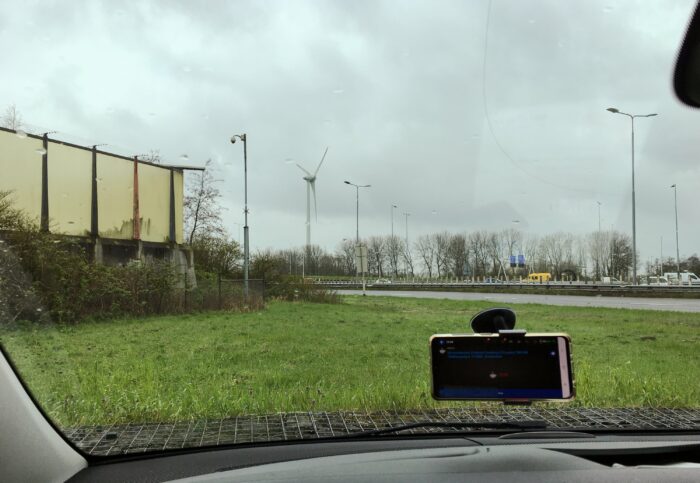 Uitzicht vanuit de voorruit van de auto. links een onduidelijk lichtgeel gebouw. Voor een grasveldje en daarachter de autosnelweg met natuurlijk wat auto's.