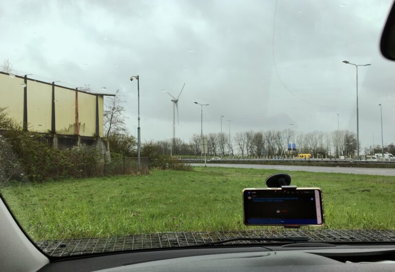 Uitzicht vanuit de voorruit van de auto. links een onduidelijk lichtgeel gebouw. Voor een grasveldje en daarachter de autosnelweg met natuurlijk wat auto's.