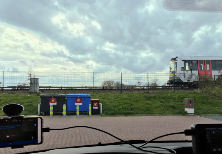 Uitzicht vanuit voorruit auto met uitzicht op het spoor van metro 53 op een sort grasdijk. Daarvoor wat containers op het parkeertereintje van het pompstation. De lucht is gevuld met dreigende wolken.