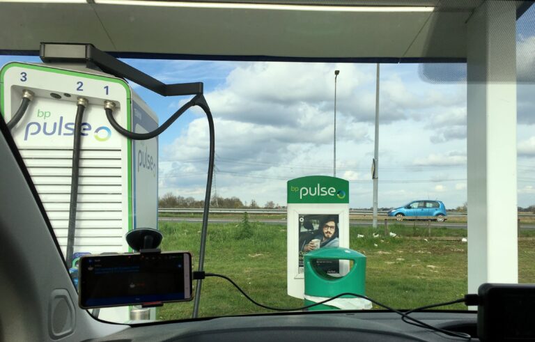 Uitzicht vanuit mijn voorruit van de auto. Je ziet een oplaadstation van pulse en een groene prullenbak en nog een reclame voor pulse. Daarachter de afrit richting Volendam met een turquoise auto. Daarachter ligt waterland, maar dat zie je slechts als een streepje aan de horizon. 