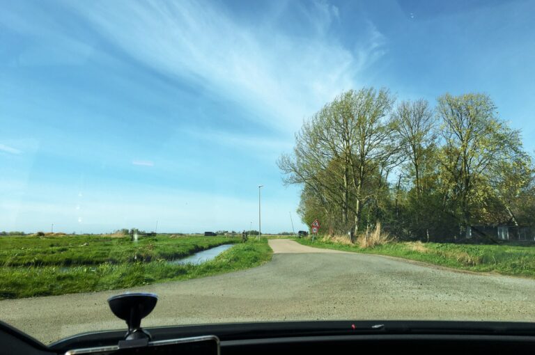 Uitzicht vanuit de voorruit van de auto op een smal weggetje met links weiland en rechts een groep bomen met een huis erin. Ook zie je deel van de sloot (die de blauwe lucht weerkaatst) aan de linkerkant van de weg.