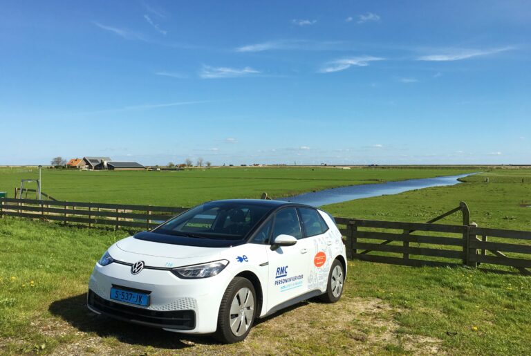 Foto van een taxi van het RMC. Een Volkswagen ID3. Geparkeerd voor een hek van een weiland met daarachter een panorama  met sloot en boerderij. Aan de horizon de Uitdammerdijk die het markermeer onder controle houdt.