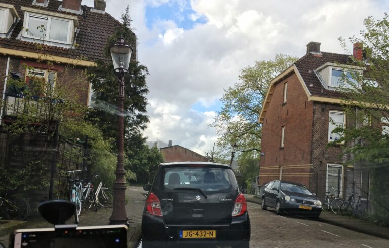 Straatje in Amsterdam Noord. Donkere auto ervoor en links en rechts jaren 50 huizen met 3 verdiepingen. Hollandse luchten op de achtergrond.