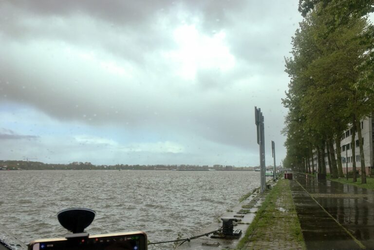 Grijzig uitzicht op een stukje van 't IJ met regenwolken. De wind maakt aardig wat golven. rechts de kade met bomen en daarachter een wit flatgebouw.