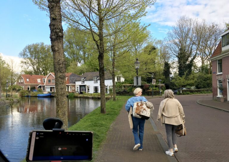 Uitzicht vanuit mijn voorruit. Aan de linkerkant van de straat geparkeerd langs het water. Tussen het water en mij een groenstrook met een paar puber bomen. Twee wandelaars op de weg en langs het water mooie pittoreske huizen.