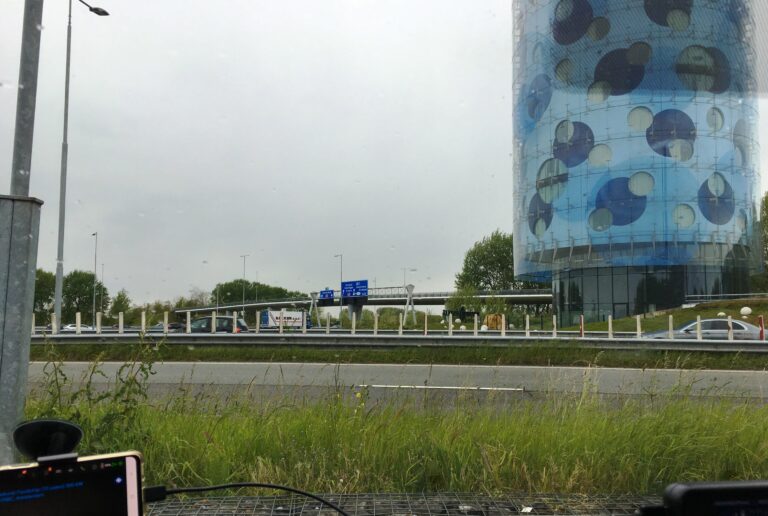 Uitzicht vanuit mijn voorruit. Zicht op klein stukje groenstrook, daarna de de snelweg en de ronde blauwe gestippelde hotel toren. Een niet te missen gebouw in Amsterdam Zuidoost.