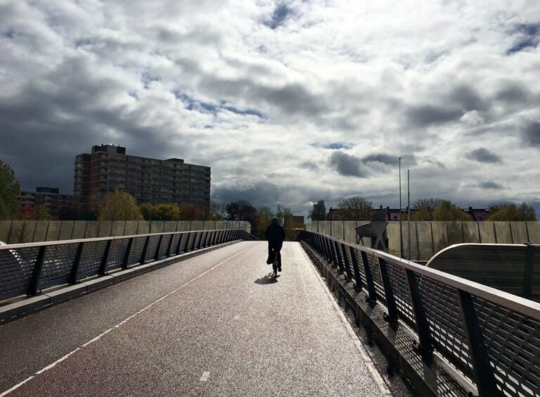 Silhouet van een fietser van achter die de bijna op de top van de fiets- en wandelbrug is die de A10 snelweg oversteekt. Dreigende wolken en een skyline van Diemen voor hem.  De zon schijnt en het natte wegdek weerkaatst 't.