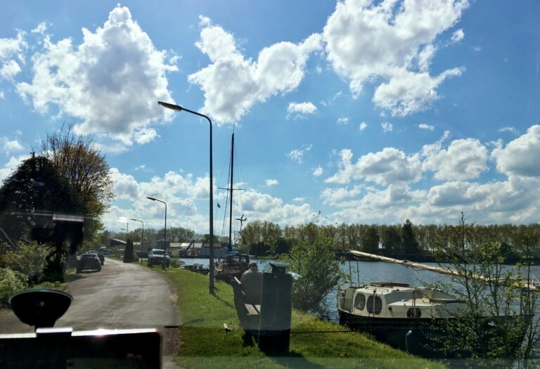 een naar links afbuigend weggetje met rechts een oever met boten en water. Op de achtergrond bomen en een heldere blauwe lucht met wat wolken. De zon schijnt volop.
