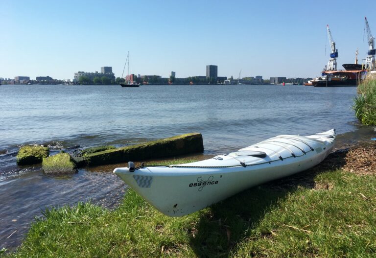 De oever van Schellingwouderpark met mijn witte kajak op het droge. Uitzicht op het IJ. Kalm water. Op de horizon skyline van Amsterdam rechts de rederij Damen en hun kranen en droogdok. Heldere blauwe lucht wat weerspiegeld in het water.
