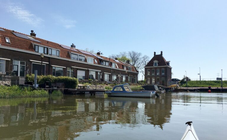 Foto vanaf het water in mijn kajak. Links een rijtje huizen langs het water met daarvoor wat boten. En nog een losstaand huis 18e-eeuws huis op de dijk.