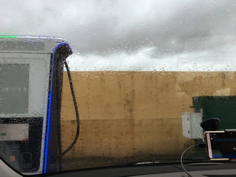 Laadpaal links, schutting van multiplex platen er achter. Daarboven nog wat blauwe lucht met witte wolken. Vertekend door de regendruppels op het voorruit.