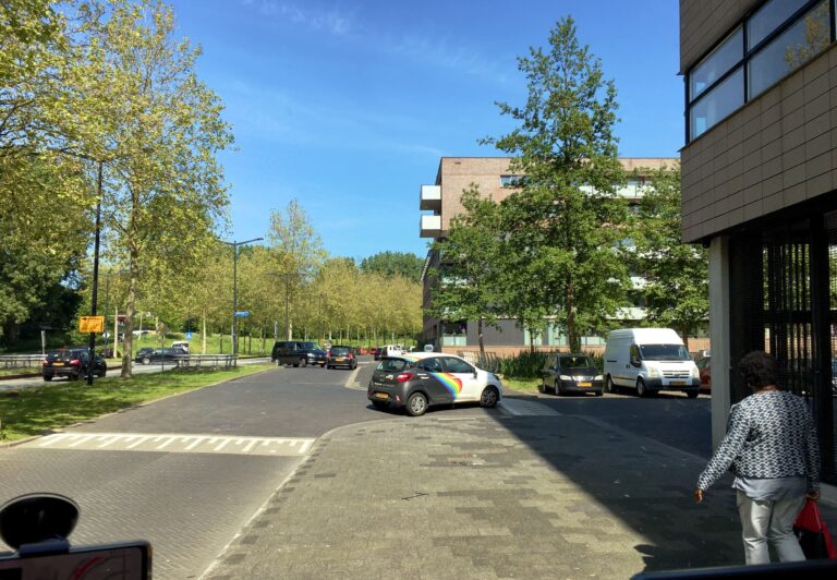 Uitzicht op een weg met links lage flats van rond de eeuwwisseling. Links bomen in het blad. Op de wegen een aantal auto's die in en uitvoegen. Rechts een vrouw die net mijn auto passeert.