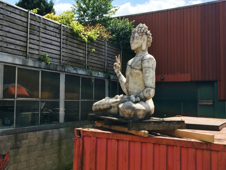 Een beeld van een soort van boeda, maar dan hipster met een v teken en een lichaam uit stukken als Frankenstein. Op een pallet met wat balkjes op weer een rode stalen scheepscontainer.