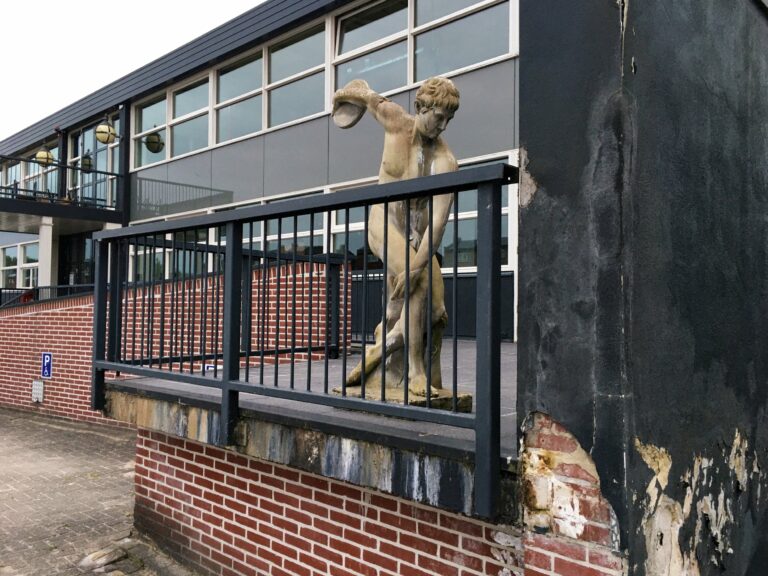 Een slechte kopie van de discuswerpen op een vergeten vervallen veranda van een semi modern kantoorgebouw. Zijn gezicht en werprichtig is naar en zwarte dikke muur die een halve meter van 'm af staat.