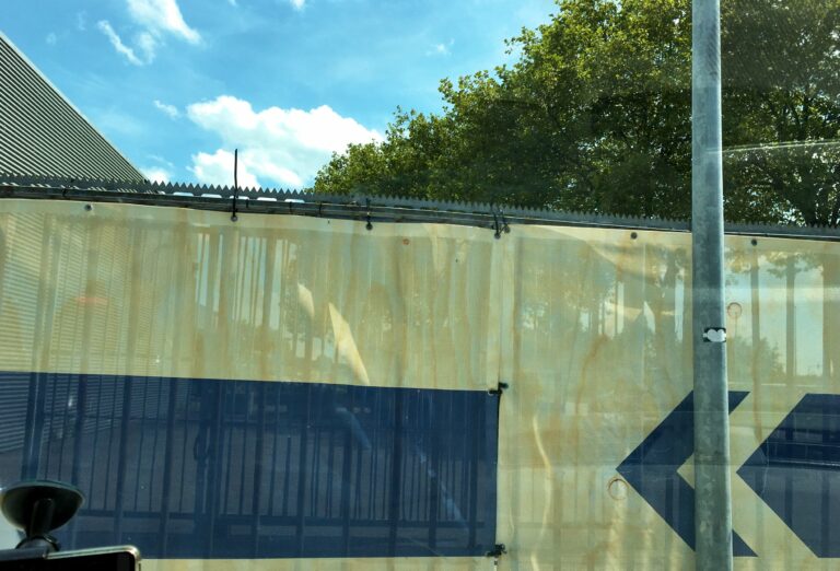 een hek met een doek van de gamma er tegenaan. Daarachter een stukje blauwe lucht en de toppen van bomen. Eigenlijk niets te zien dus.