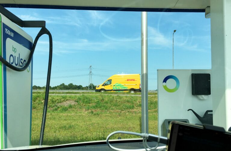 Doorkijkje bij een oplaadpunt van bp. Links de laadpaal en daaromeen de constructie. In het doorkijkje een stuk gras en daarachter een weg met een geel DHL busje. Een mooie blauwe lucht als achtergrnd.