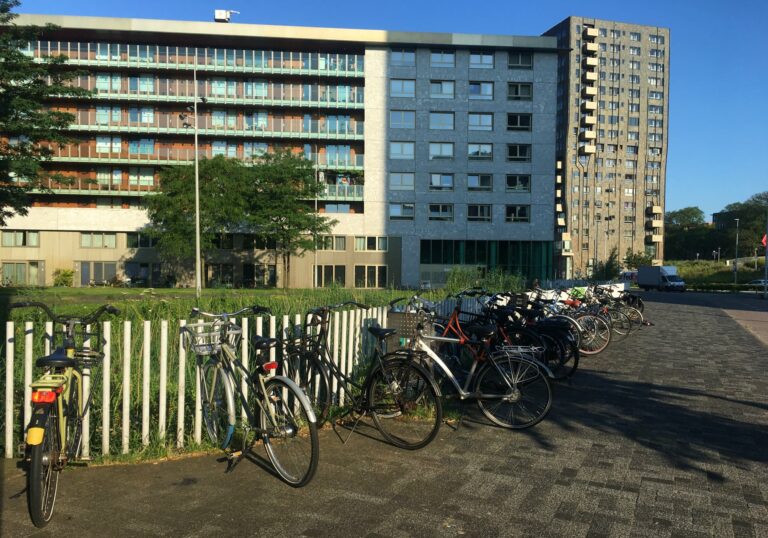 Fietsen tussen de spijlen van een wit hek geplaatst. Op de achtergrond witte flatgebouwen en een helder blauwe lucht