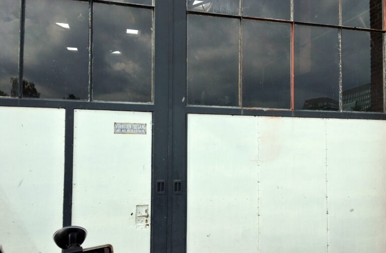 Een garagedeur. Aan de onderkant wit. Daarboven glas waar de wolken in spiegelen. Het kozijn is zwart en er staat een "verboden toegang" bordje op.