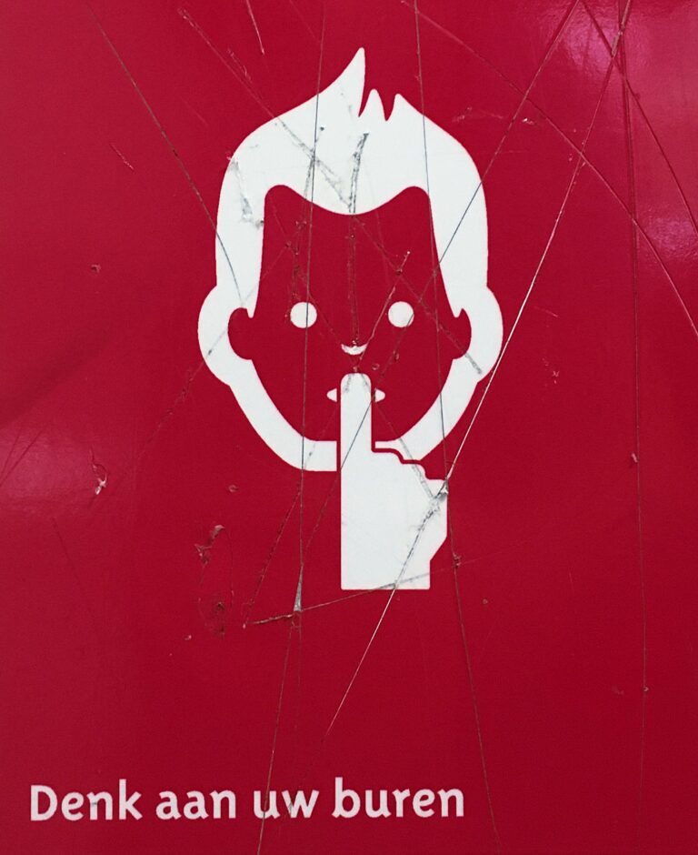 Waarschuwingsbord in diep rood met een wit pictogram van een hoofd met "vlammend" haar en een vinger op de lippen. Linksonder de tekst "Denk aan uw buren"
