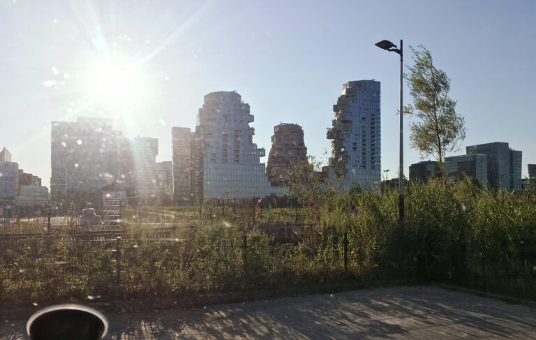 Een panorama van de zuidas met hoge moderne flatgebouwen. Meest prominent is degene waar het lijkt of het middelste deel is ingestort. Op de voorgrond de parkeervakken waar ik sta geparkeerd met wat groen begroeiing. Tussen de flatgebouwen en mij zitten sportvelden. 