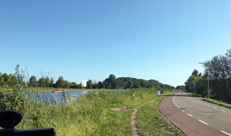 Ik sta aan de dijk langs de Amstel links van mij. Rechts de weg de naar de horizon loopt. Op het water een oranje boot. Aan de andere oever zijn volwassen bomen zichtbaar. De lucht is wolkloos blauw.