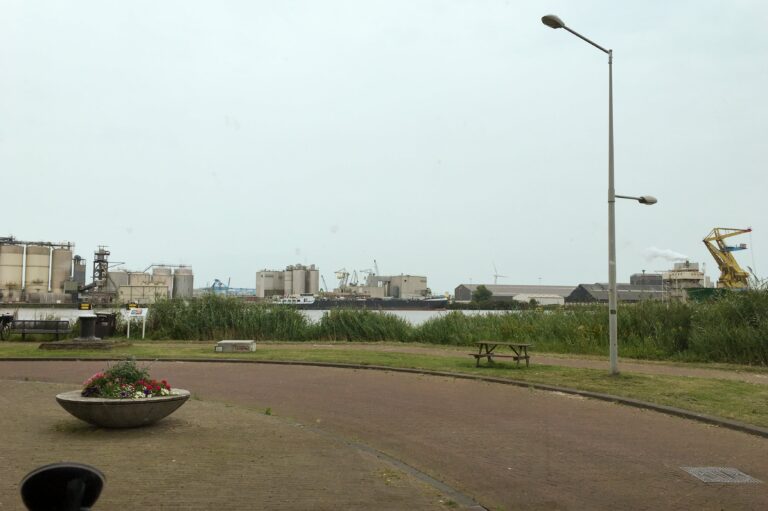 Sta links geparkeerd langs een smalle weg met een bocht naar links. Aan de rechterkant is een begroeide oever. Aan de overkant van 't IJ is zij de gebouwen van de havenindustrie te zien. Inclusief kranen.