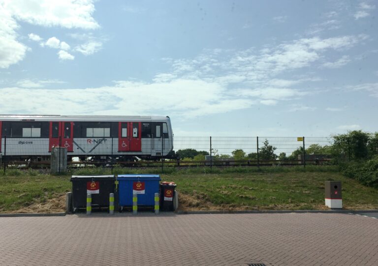 Uitzicht vanuit autoraam bij tankstation. Er is wat bestrating en links staan twee afvalcontainers. Daarachter een graswal en een hek met het spoor van de metro. De metro komt er net aan van links en is halverwege de foto. De lucht is blauw met wat sluierbewolking.