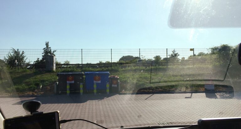 uitzicht vanuit autoraam bij tankstation. Je ziet een stukje straat wat overgaat in een graswal met daarop een hek. Daarachter (onzichtbaar) loopt het spoor van de metro. Links voor het gras twee afvalcontainers. We hebben een blauwe licht.