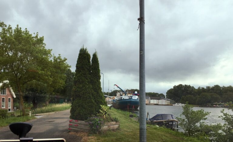 Sta links langs de weg geparkeerd aan de groene oever. Rechts water Smal Weesp met wat boten langs de kant op de achtergrond een fabriek en bomen. Links een vrijstaand huis en veel bomen. Recht voor me twee coniferen. De lucht is bewolkt.