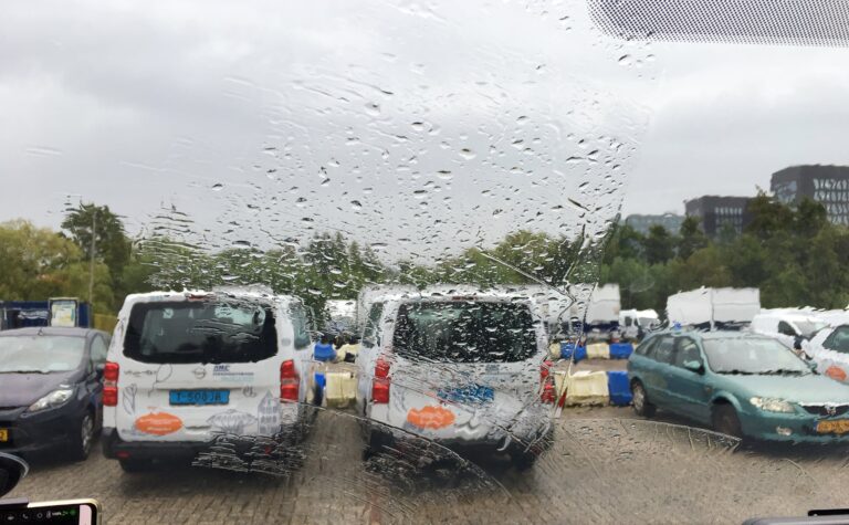 Uitzicht op een parkeerplaats. Het raam is bedekt met water en dat vertekend de auto's die er staan. Op de achtergrond bomen en rechts daarachter een paar kantoorgebouwen
