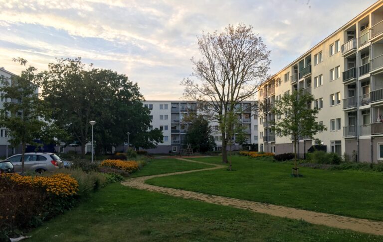 Een mooi binnenplaatsje (gras en wat bomen) tussen 3 appartementencomplexen in. Ze staan haaks op elkaar en 5 hoog. Links ook nog wat parkeergelegenheid en 2 auto's. De zon is aan het ondergaan en schijnt oranje licht op de bvenkant van het gebouw rechts. Ook de wolken kleuren een beetje oranje.