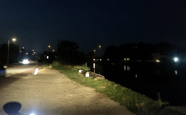Het is donker. Ik sta langs het water aan de oever op een zanderige parkeerplaats. Strookje groen is de grens tussen het water rechts en mij. Links de weg met wat auto's. Ik verlicht alles met mijn koplampen. De rest zijn wat lichtjes aan de horizon met de water als een spiegel.