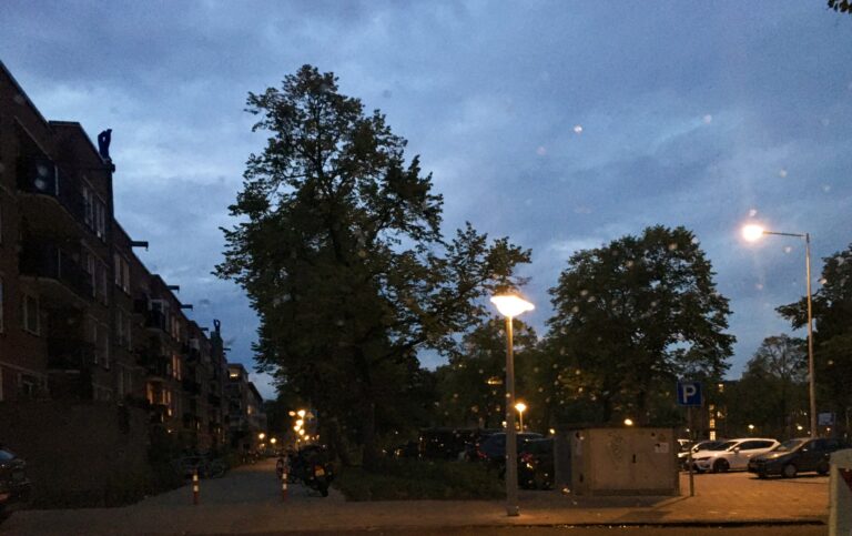 Uitzicht vanuit de auto tijdens mijn pauze. Tijdens de schemeruren. Links een siloët van een  appartementencomplex uit de jaren 80 van 4 hoog. in het midden wat grote bomen en een bewolkte lucht. recht een parkeerplaats met auto's